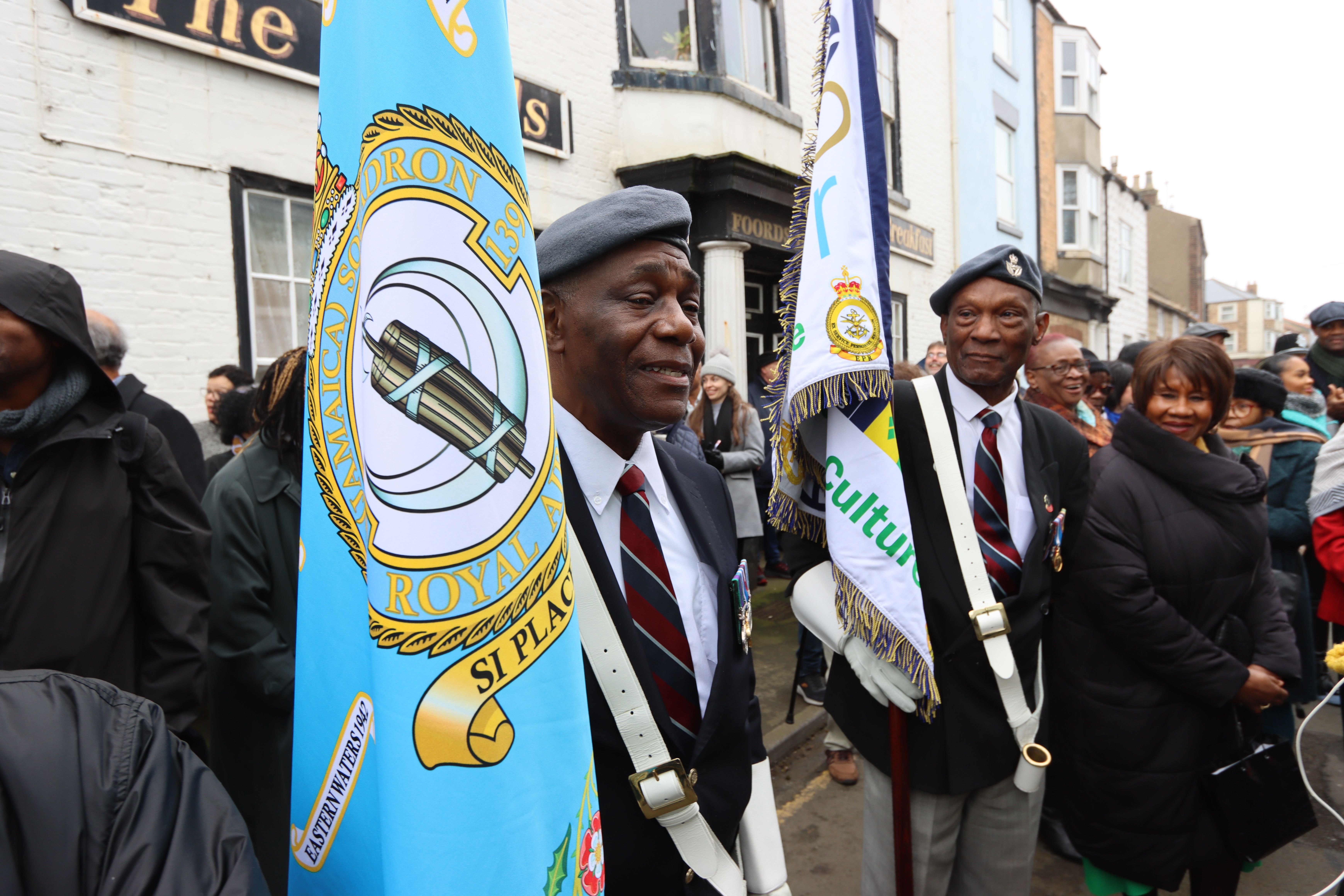 Image shows RAF Veterans carrying Squadron colour flag with civilians in the streets.